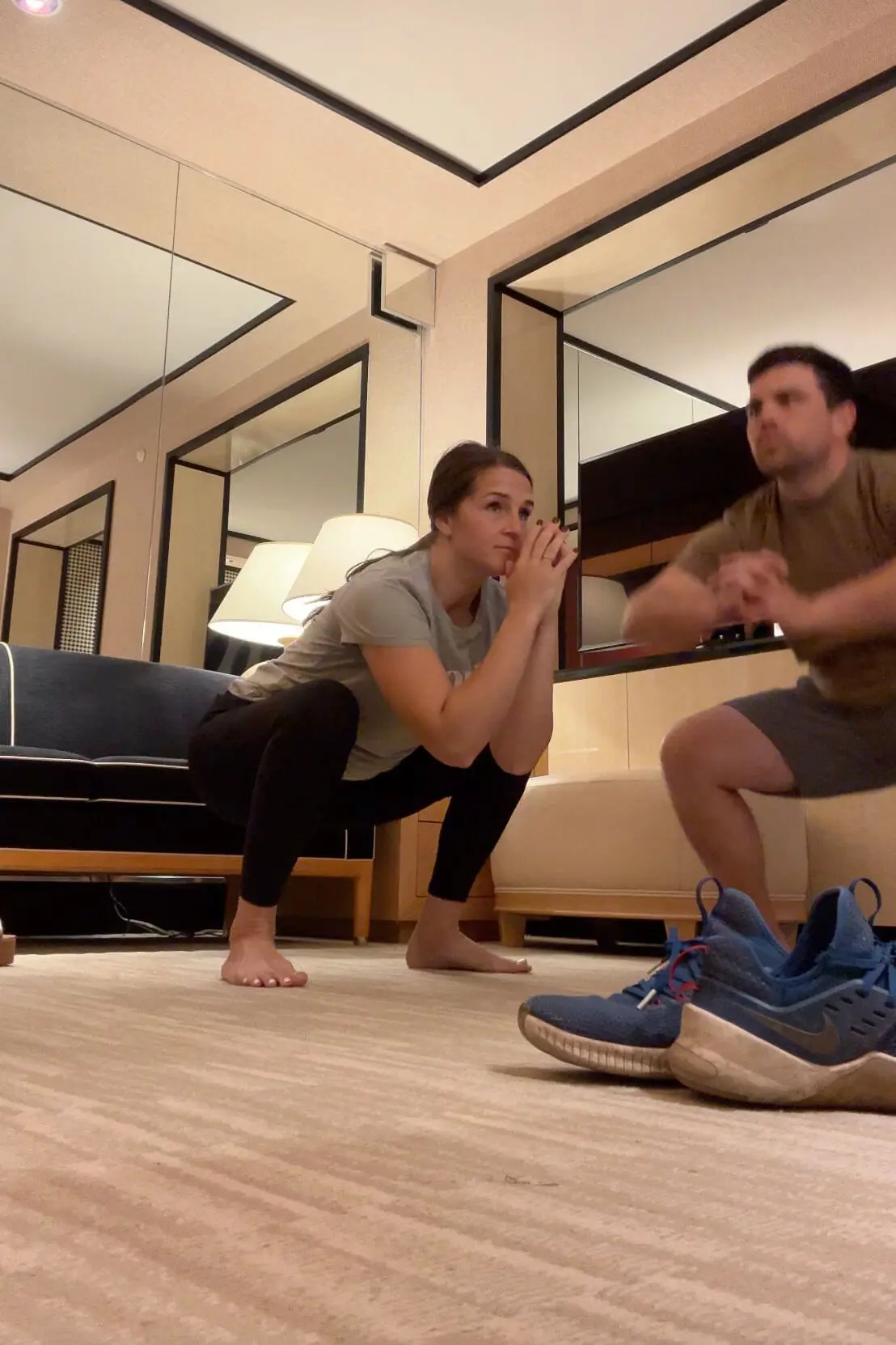 Woman and man performing squats together as part of an indoor workout routine in a well-lit living room. Both focused and in sync, they demonstrate the power of a supportive workout routine that can be done at home.